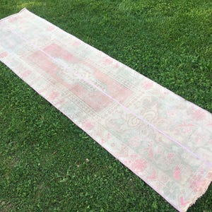 May include: A long, rectangular pink and white runner rug with a floral pattern. The rug is laid out on a green grassy surface.
