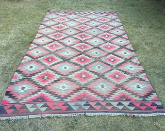 May include: A traditional Turkish kilim rug with a geometric pattern in shades of red, pink, brown, and gray. The rug has a fringed edge.