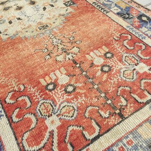 May include: A vintage rug with a red, cream, and blue geometric pattern. The rug has a worn, distressed look and features a central floral design.