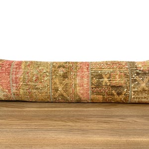 May include: A long, rectangular pillow with a patchwork design. The pillow features a mix of colors, including red, green, and tan. The pillow is made of a textured fabric, and it appears to be a decorative item for a bed or sofa.