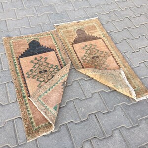 May include: Two beige prayer rugs with geometric patterns in shades of green, brown, and black. The rugs are folded and lying on a grey concrete surface.