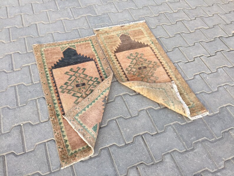 May include: Two beige prayer rugs with geometric patterns in shades of green, brown, and black. The rugs are folded and lying on a grey concrete surface.