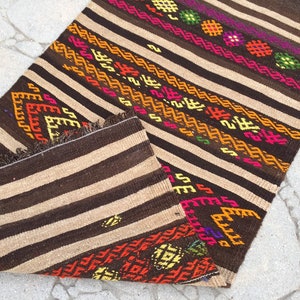 Puede incluir: Una alfombra de rayas marrones y beige con patrones geom&eacute;tricos coloridos. La alfombra tiene flecos en los bordes y est&aacute; doblada por la mitad.