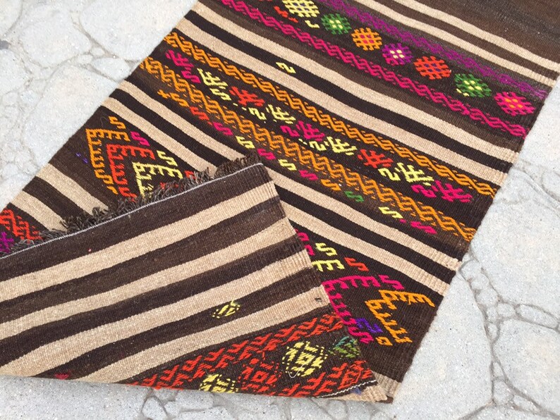 Puede incluir: Una alfombra de rayas marrones y beige con patrones geom&eacute;tricos coloridos. La alfombra tiene flecos en los bordes y est&aacute; doblada por la mitad.