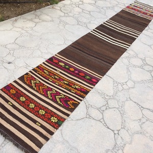 Puede incluir: Una alfombra larga, de rayas marrones y blancas con coloridos patrones geom&eacute;tricos tejidos en el dise&ntilde;o. La alfombra est&aacute; colocada sobre una superficie de hormig&oacute;n gris.