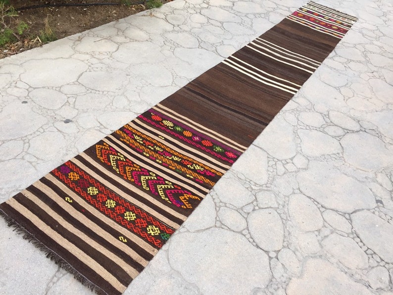 Puede incluir: Una alfombra larga, de rayas marrones y blancas con coloridos patrones geom&eacute;tricos tejidos en el dise&ntilde;o. La alfombra est&aacute; colocada sobre una superficie de hormig&oacute;n gris.