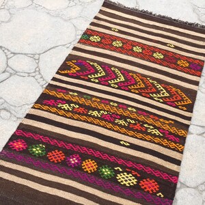 Puede incluir: Una alfombra de rayas marrones y beige con patrones geom&eacute;tricos coloridos. La alfombra tiene flecos en el borde superior.