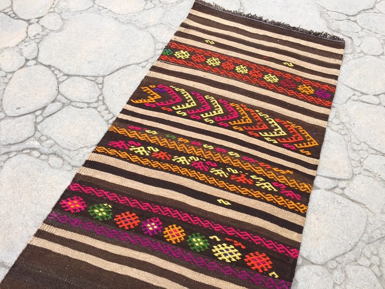 Puede incluir: Una alfombra de rayas marrones y beige con patrones geom&eacute;tricos coloridos. La alfombra tiene flecos en el borde superior.