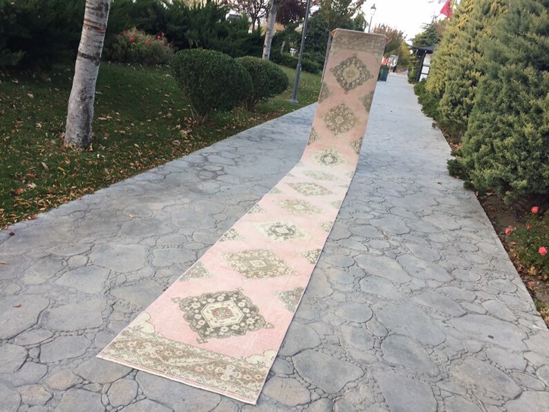 May include: A long, pink and cream runner rug with a geometric pattern. The rug is laid out on a gray concrete walkway.