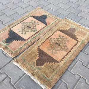 May include: Two small, rectangular, vintage rugs with a geometric pattern in shades of brown, green, and pink. The rugs have a fringed border.