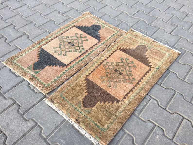 May include: Two small, rectangular, vintage rugs with a geometric pattern in shades of brown, green, and pink. The rugs have a fringed border.