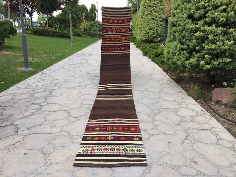 Puede incluir: Una alfombra larga y estrecha de color marr&oacute;n y blanco con patrones geom&eacute;tricos coloridos tejidos en el dise&ntilde;o. La alfombra est&aacute; colocada en un camino de piedra.
