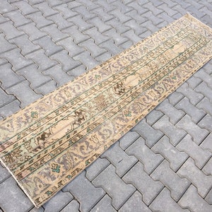 May include: A long, narrow beige rug with a floral pattern in shades of brown, green, and pink. The rug has a worn, vintage look and is laid out on a gray concrete surface.