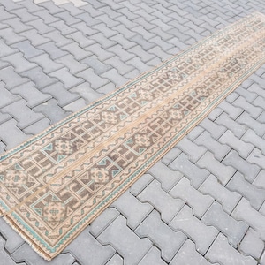 Può includere: Un tappeto lungo e rettangolare con un motivo geometrico in tonalità di marrone, beige e turchese. Il tappeto ha un bordo con frange ed è steso su una superficie di cemento grigia.