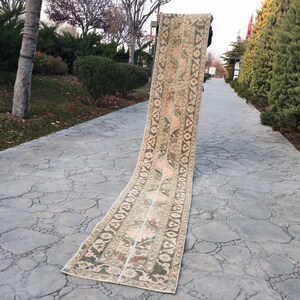 Puede incluir: Una alfombra larga y estrecha con fondo beige y intrincados patrones florales en tonos verdes, rosas y marrones. La alfombra está colocada sobre un camino gris y texturizado.