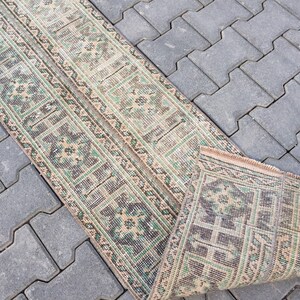May include: A beige and green patterned runner rug with a geometric design. The rug is rolled up and lying on a gray concrete surface.