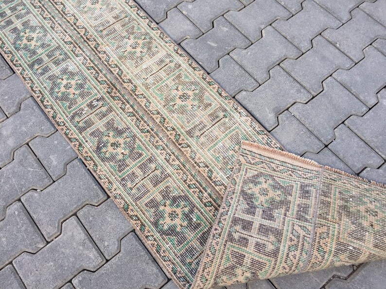 May include: A beige and green patterned runner rug with a geometric design. The rug is rolled up and lying on a gray concrete surface.