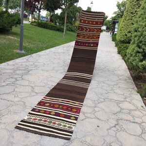 Puede incluir: Una alfombra larga, marr&oacute;n y blanca tejida con patrones geom&eacute;tricos coloridos. La alfombra est&aacute; colocada en un camino de piedra.