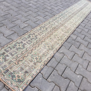 Puede incluir: Una alfombra larga y rectangular con fondo beige y un patrón geométrico verde y marrón. La alfombra está sobre una superficie de hormigón gris.