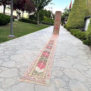 May include: A long, narrow runner rug featuring a repeating pattern of circular medallions in shades of deep pink, olive green, and beige. The rug is laid out flat on a stone pathway, with one end standing upright to display the intricate textile design.