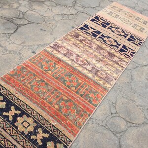 May include: A long, narrow rug with a colorful, geometric pattern. The rug is made of woven fabric and has a distressed, vintage look. The rug is made up of several different panels, each with a unique design. The colors in the rug include black, red, orange, green, and beige.