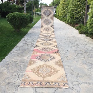 May include: A long, narrow, cream-coloured rug with a geometric pattern in shades of brown, pink, and blue. The rug is laid out on a stone path.