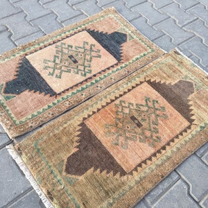 May include: Two small, rectangular rugs with a geometric pattern in shades of brown, green, and pink. The rugs have a fringed border.