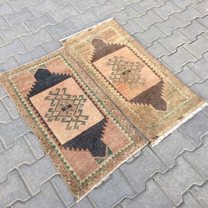 May include: Two small, rectangular, hand-woven rugs with a geometric pattern in shades of brown, green, and beige. The rugs have a fringe border.