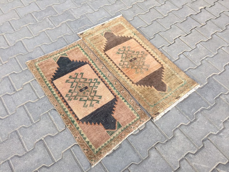 May include: Two small, rectangular, hand-woven rugs with a geometric pattern in shades of brown, green, and beige. The rugs have a fringe border.