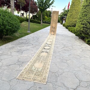 May include: A long, narrow runner rug featuring a beige and cream base with intricate, symmetrical dark patterns and geometric motifs. The rug is displayed vertically, extending along a stone pathway, showcasing a detailed, repeating design.