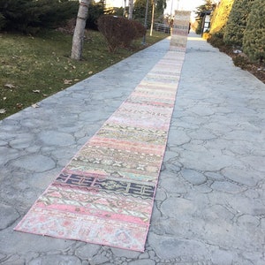 Puede incluir: Una alfombra vintage larga y estrecha, de color rosa y multicolor con un patrón geométrico, colocada sobre un camino de concreto gris.