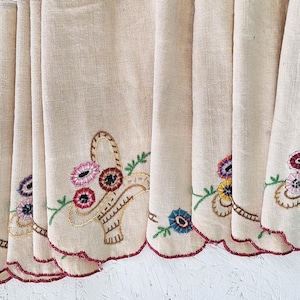 May include: A set of eight beige linen napkins featuring scalloped edges with dark red stitching. Each napkin displays an embroidered basket motif filled with colorful flowers in shades of pink, blue, and yellow, accented with green leaves.