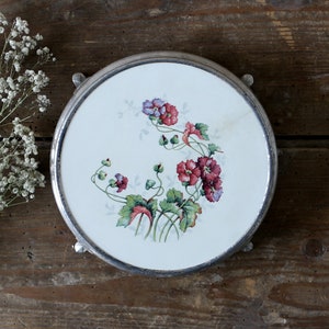 Peut inclure: Un dessous de plat rond doté d'un centre en céramique blanche orné d'une illustration florale de fleurs rouges et violettes avec des feuilles vertes, encadré par un contour en métal argenté muni de petits pieds. Le motif est détaillé.