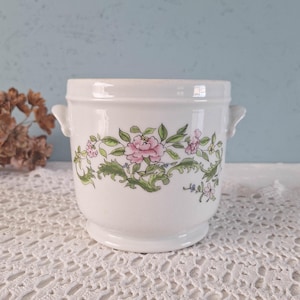 May include: A white ceramic pot with two small side handles, featuring a decorative band of hand-painted pink flowers and green leaves encircling the centre. The vessel has a smooth, glossy finish, a flared rim, and a raised circular base.