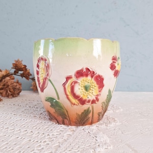 May include: A ceramic planter with a scalloped rim featuring a gradient finish that transitions from pale green at the top to a warm peach hue at the base. The exterior is decorated with embossed red and yellow poppy flowers and green leaves.