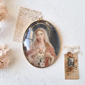 Puede incluir: Imagen religiosa ovalada en un marco dorado. La imagen representa a una mujer con un halo, vestida con una túnica rosa y un velo blanco, con un corazón y flores en el pecho. También hay una pequeña etiqueta con una cinta y una nota manuscrita.