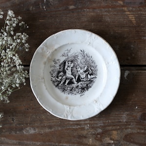 May include: A white ceramic plate with a black and white design of three cherubs playing in a garden. The plate has a raised rim and a scalloped edge.