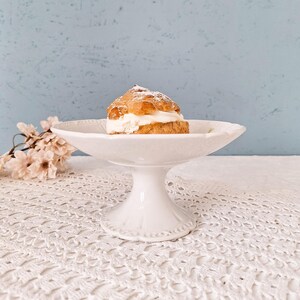 Puede incluir: Un cuenco de cerámica blanca sobre pedestal con borde festoneado y detalles en relieve, que contiene un profiterol espolvoreado con azúcar glas. El cuenco presenta una base acampanada y un acabado liso y brillante de diseño clásico.
