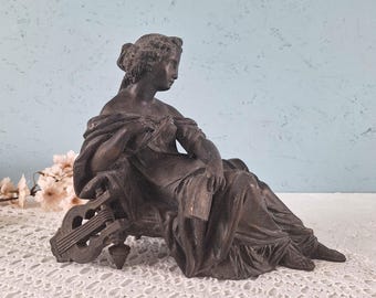 Könnte beinhalten: Eine dunkel bronzefarbene Skulptur einer sitzenden Figur in fließenden Gewändern, die eine Hand auf eine kleine Leier stützt. Die Figur hat eine detaillierte Frisur und hält einen rechteckigen Gegenstand in der anderen Hand, mit drapiertem Gewand.