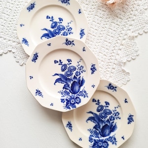 Puede incluir: Tres platos de cerámica blanquecina con un diseño floral azul. Cada plato presenta una disposición diferente de flores y hojas azules. Los platos están colocados sobre una superficie de encaje blanco, con flores rosas en el fondo.