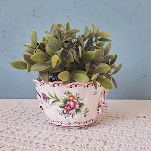 May include: A white ceramic planter with a scalloped rim and ornate handles, featuring a hand-painted floral motif with pink, red, and green accents. The planter contains a small, leafy green artificial plant with multiple stems and foliage.