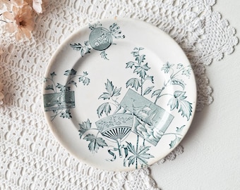 May include: A round ceramic plate featuring a white base with a dark teal transferware pattern. The design includes botanical leaf motifs, a handheld folding fan, and rectangular vignettes depicting sailing ships on the water.