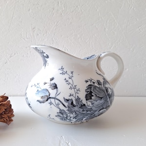 Puede incluir: Jarra de cerámica blanca con cuerpo redondeado y asa. Está decorada con un diseño floral azul y gris. El pico es curvo y el asa es un simple lazo. Un objeto decorativo.