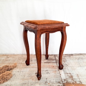 Peut inclure: Un petit tabouret en bois antique avec une assise carr&eacute;e et rembourr&eacute;e. Le tabouret a quatre pieds incurv&eacute;s et est en bois brun fonc&eacute;. Le coussin de si&egrave;ge est de couleur brun-orang&eacute; chaud. Le tabouret mesure environ 46 cm de haut.