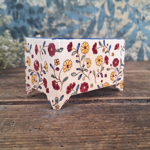 Könnte beinhalten: Kleiner, rechteckiger Keramik-Pflanztopf mit floralem Muster. Der Pflanztopf hat eine cremefarbene Basis, die mit roten und gelben Blumen, grünen Blättern und einem blauen Rand verziert ist.