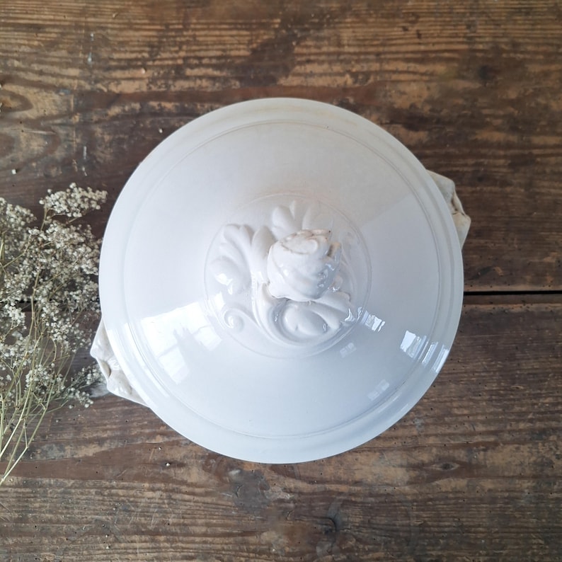 May include: A white ceramic serving dish lid with a raised floral design in the center. The lid is round with a slightly domed shape and a glossy finish. The background is a rustic wooden surface.