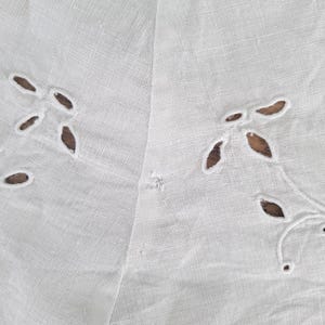 Puede incluir: Un tejido blanco que presenta un delicado patr&oacute;n de bordado ingl&eacute;s con peque&ntilde;os recortes en forma de hoja dispuestos en grupos. El material tiene una textura tejida visible y una calidad ligera y semitransparente con una peque&ntilde;a imperfecci&oacute;n.