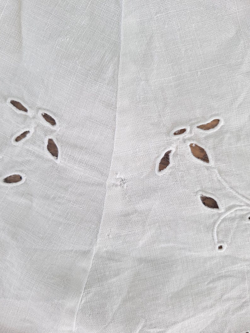 Puede incluir: Un tejido blanco que presenta un delicado patr&oacute;n de bordado ingl&eacute;s con peque&ntilde;os recortes en forma de hoja dispuestos en grupos. El material tiene una textura tejida visible y una calidad ligera y semitransparente con una peque&ntilde;a imperfecci&oacute;n.