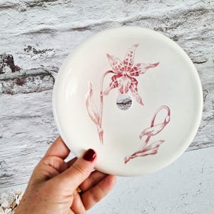 Könnte beinhalten: Eine runde, cremeweiße Seifenschale aus Keramik mit einem zentralen Abflussloch und einer zarten, rot-rosa botanischen Illustration einer lilienartigen Blume mit Stängeln und Blättern auf der Oberfläche der Schale.