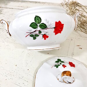 May include: White porcelain serving bowl with a lid. The bowl and lid feature a red rose with green leaves and gold trim.